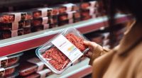 Above the shoulder view of woman holding a pack of beef mince in a supermarket. Standing in front of the meat product aisle and have no idea which product to choose from. The Conscious Carnivore - find meat that is locally-sourced, healthy, high quality butchering, and handled with respect. The environmentally conscious way to choosing food.