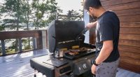 Man grilling meat and vegetables on a patio overlooking a lake.