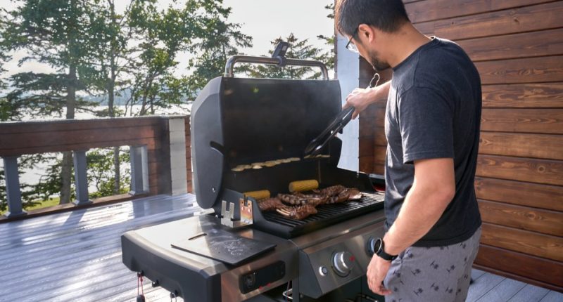 Man grilling meat and vegetables on a patio overlooking a lake.