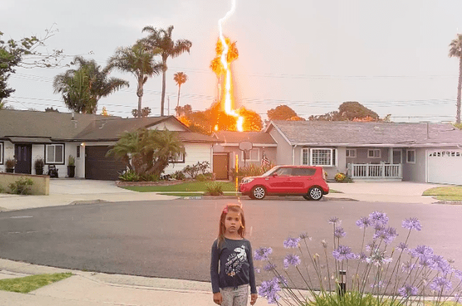 Lightning striking a palm tree near a house with a young girl standing nearby.