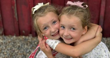 Twin girls hugging in front of a red wooden wall.
