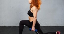 Young redhead woman performing frontal lunge with the dumbbells at the gym