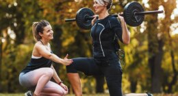 A mature woman is doing lunges exercises with personal trainer in the park, dressed in a black suit with an EMS electronic simulator to stimulate her muscles.