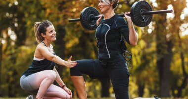 A mature woman is doing lunges exercises with personal trainer in the park, dressed in a black suit with an EMS electronic simulator to stimulate her muscles.