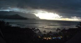 Ocean view at sunset with dark clouds and mountains in the background.