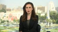 News anchor sitting in front of a window overlooking a city.