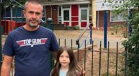 A girl and her father stand near floral tributes outside a school.