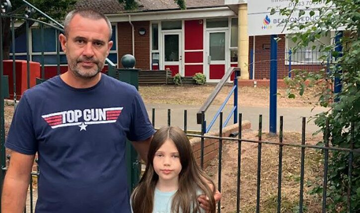 A girl and her father stand near floral tributes outside a school.