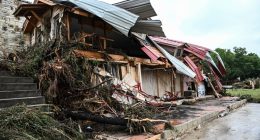 Flood-damaged home with roof torn off and debris scattered around.