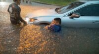 Two men help a person out of a car submerged in floodwaters.