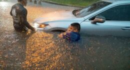 Two men help a person out of a car submerged in floodwaters.