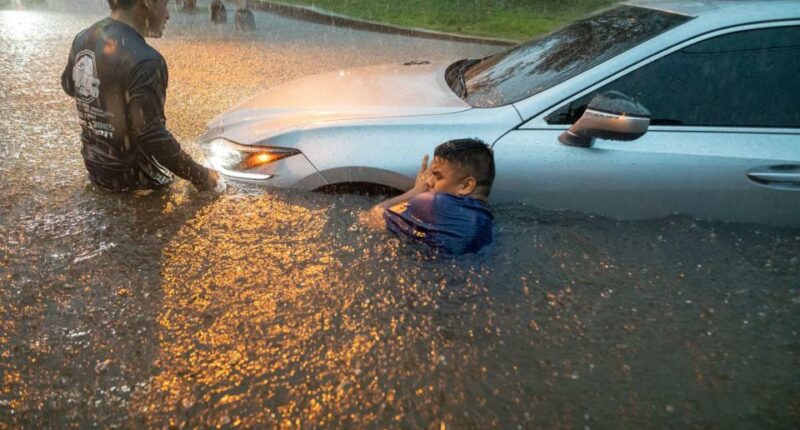 Two men help a person out of a car submerged in floodwaters.