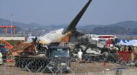 Burned wreckage of a plane at an airport with emergency personnel on the scene.