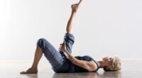 Single athletic woman with blond hair in blue outfit on back stretching hamstring muscles for leg in mid air