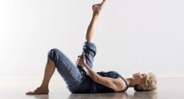 Single athletic woman with blond hair in blue outfit on back stretching hamstring muscles for leg in mid air