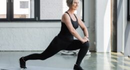 full length of happy senior woman in sportswear doing lunges on fitness mat near dumbbells in gym