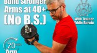 A man with gray hair and glasses exercises by lifting a dumbbell, showcasing strength and determination in a vibrant blue gym environment. Building Stronger Arms After 40 Could Be Way Easier Than You