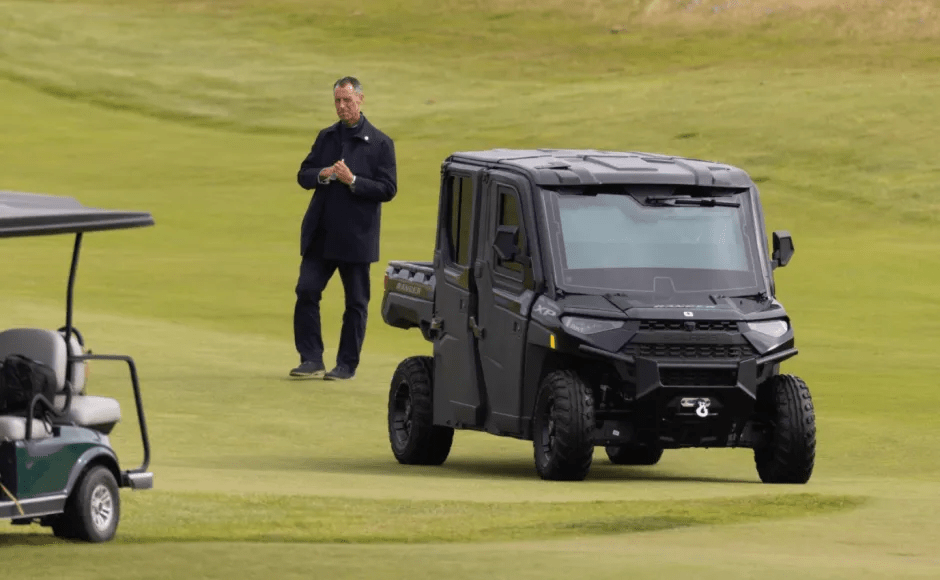 Man standing on a golf course near a golf cart and a black utility vehicle.