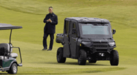 Man standing on a golf course near a golf cart and a black utility vehicle.