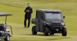 Man standing on a golf course near a golf cart and a black utility vehicle.