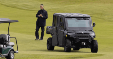 Man standing on a golf course near a golf cart and a black utility vehicle.