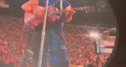 Chris Martin playing guitar on a large screen at a concert.