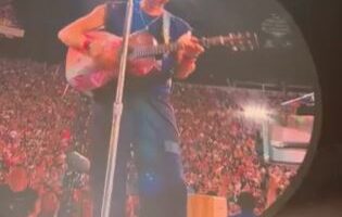 Chris Martin playing guitar on a large screen at a concert.