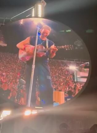 Chris Martin playing guitar on a large screen at a concert.