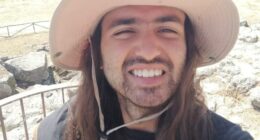 Man with long hair and a hat smiling at a landscape.