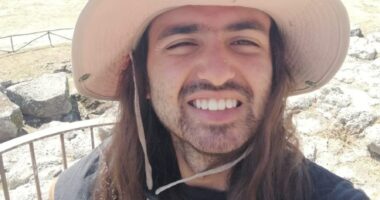 Man with long hair and a hat smiling at a landscape.
