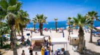 Crowded beach in Palaio Faliro, Athens, with palm trees and beach umbrellas.