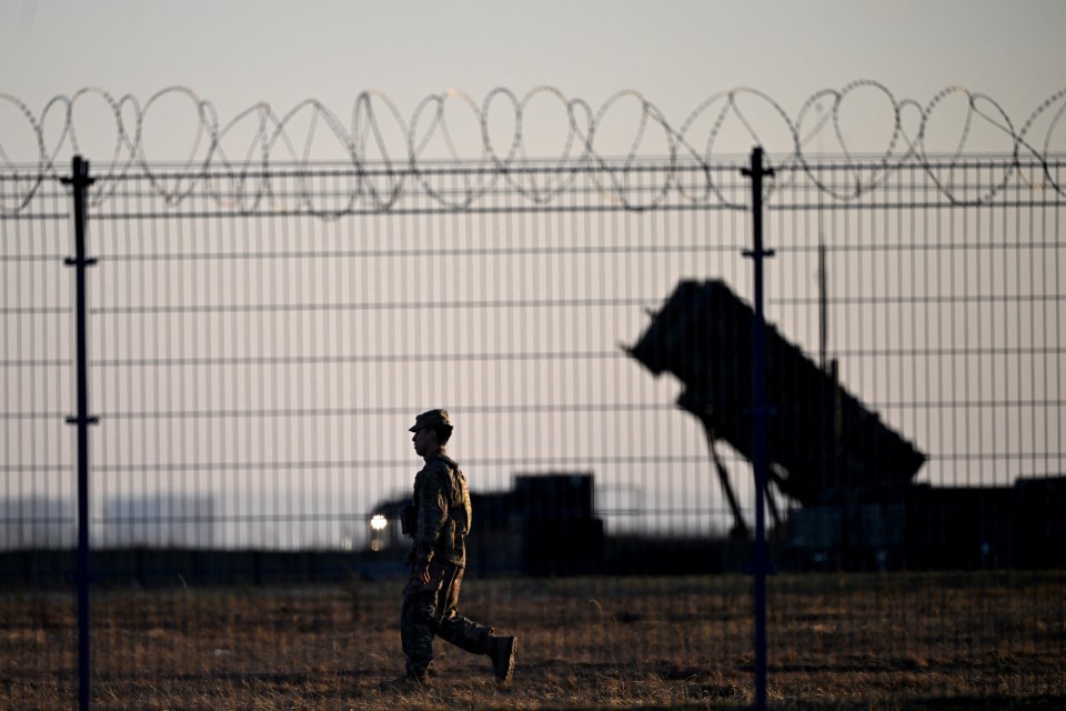Soldier walking past Patriot air defense systems at a military hub.