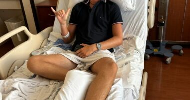 Teenager in hospital bed with bandaged foot, giving shaka sign.