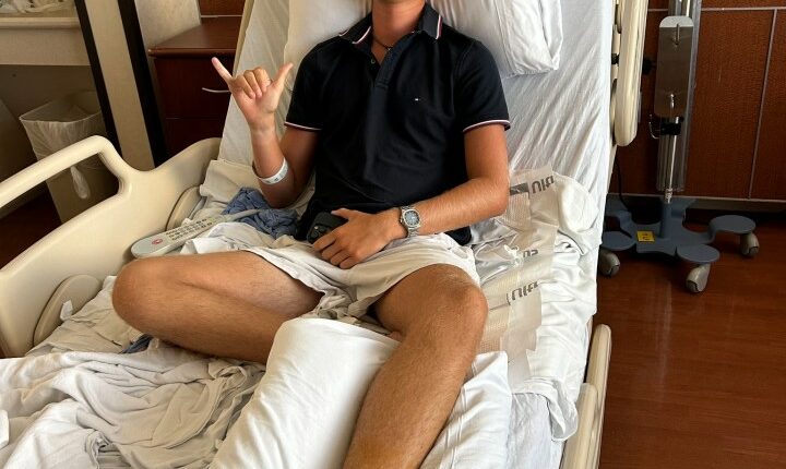 Teenager in hospital bed with bandaged foot, giving shaka sign.