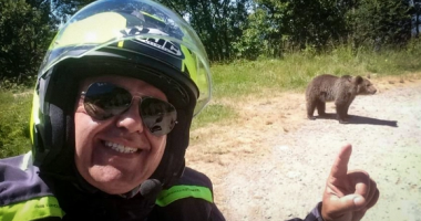 A motorcyclist smiling and pointing at a bear on a road.