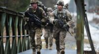 Two soldiers carrying rifles walk across a metal bridge.