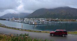A red SUV drives past a harbor filled with fishing boats.