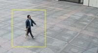 Security camera image of a man carrying a rifle walking in front of a building.