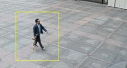 Security camera image of a man carrying a rifle walking in front of a building.