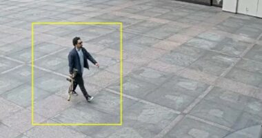 Security camera image of a man carrying a rifle walking in front of a building.