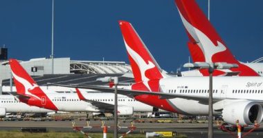 The vertical stabilisers of Qantas planes at Sydney Kingsford-Smith Airport, from the left: a Boeing B737-838 plane, registration VH-XZD; a Boeing B787-9 plane, registration VH-ZNE; and an Airbus A380-842 plane, registration VH-OQB. In the background on the left is a fourth Qantas plane, a Boeing B737-838, registration VH-XZH. In the far background is the international terminal.  In the foreground are navigational light structures at the northern end of the runway.  This image was taken from Nig