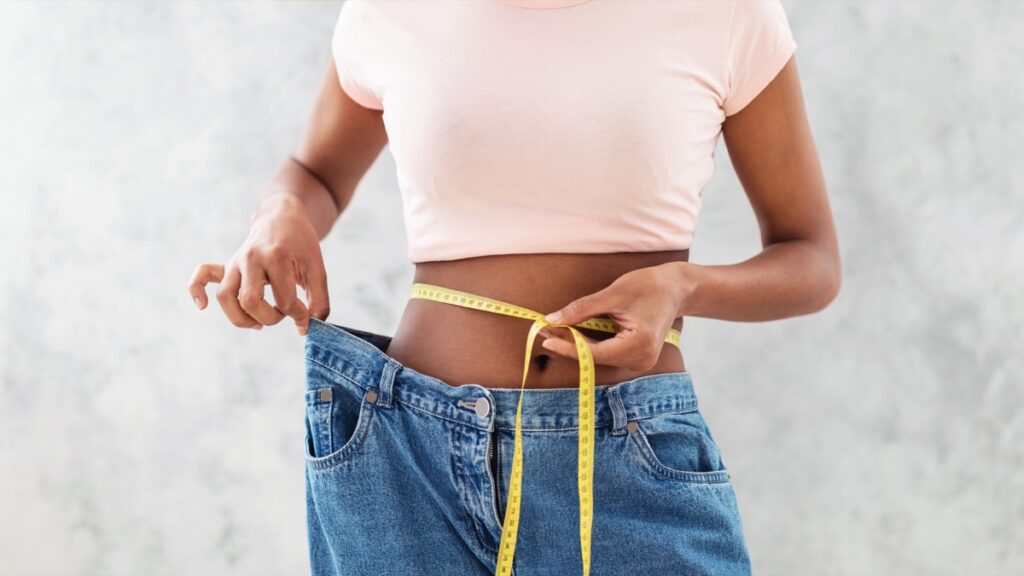 Closeup of black woman in old big jeans measuring her waist with tape, showing results of slimming diet or liposuction, grey background. Young lady promoting healthy nutrition for weight loss