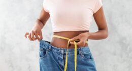 Closeup of black woman in old big jeans measuring her waist with tape, showing results of slimming diet or liposuction, grey background. Young lady promoting healthy nutrition for weight loss
