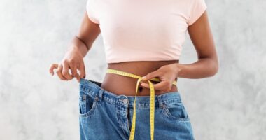 Closeup of black woman in old big jeans measuring her waist with tape, showing results of slimming diet or liposuction, grey background. Young lady promoting healthy nutrition for weight loss