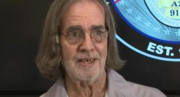 Man with long gray hair and glasses speaks in front of a police department emblem.