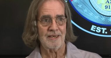 Man with long gray hair and glasses speaks in front of a police department emblem.