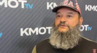 A man with a long gray beard and a USA hat speaks in front of a KMOT TV backdrop.