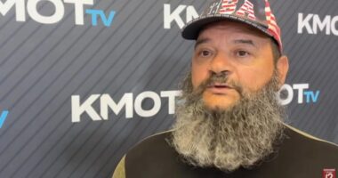 A man with a long gray beard and a USA hat speaks in front of a KMOT TV backdrop.