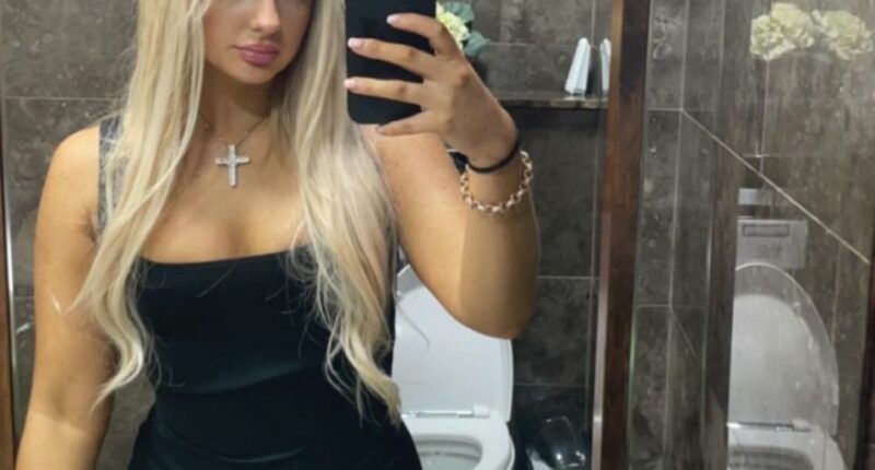 Selfie of a blonde woman in a black dress in a restroom.