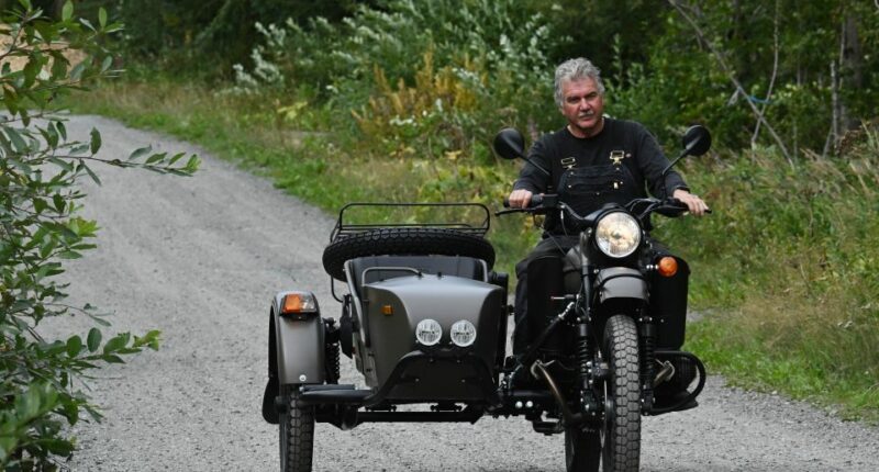 Man riding a Ural motorcycle with a sidecar on a gravel road.